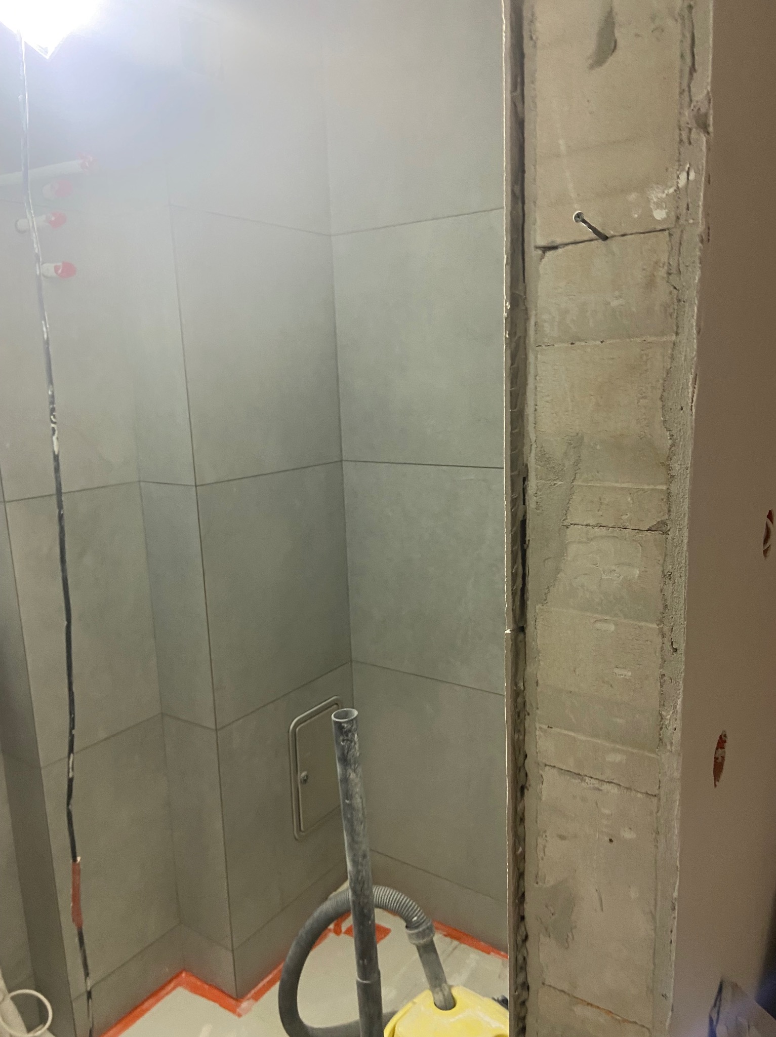 Szare płytki w rogu łazienki, widoczny fragment futryny z surowego betonu, żółty odkurzacz. Prace wykończeniowe w trakcie realizacji.