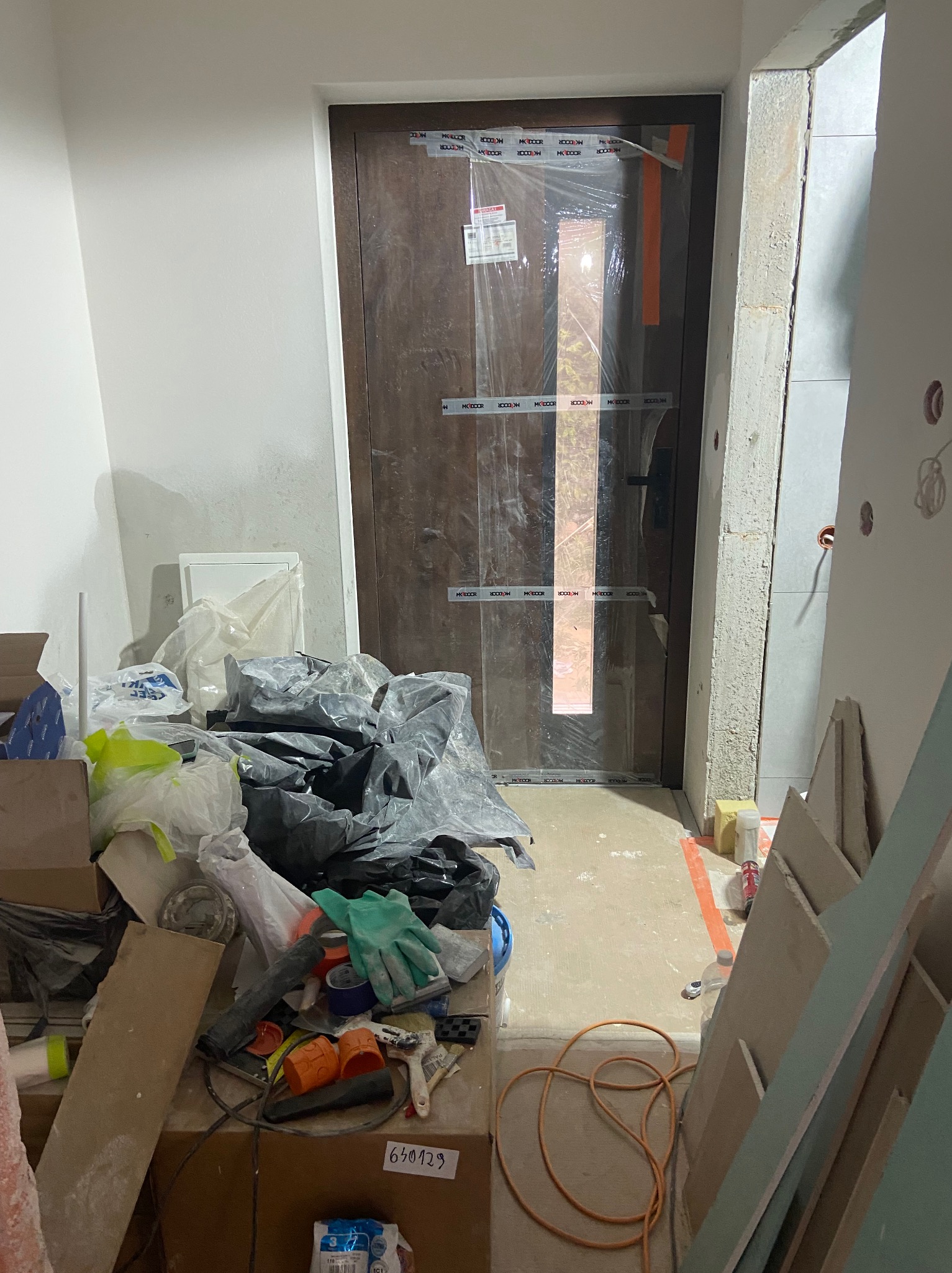 Montaż drzwi wewnętrznych w trakcie remontu: widoczne zabezpieczone folią skrzydło, surowa ościeżnica, narzędzia i materiały budowlane w pomieszczeniu.