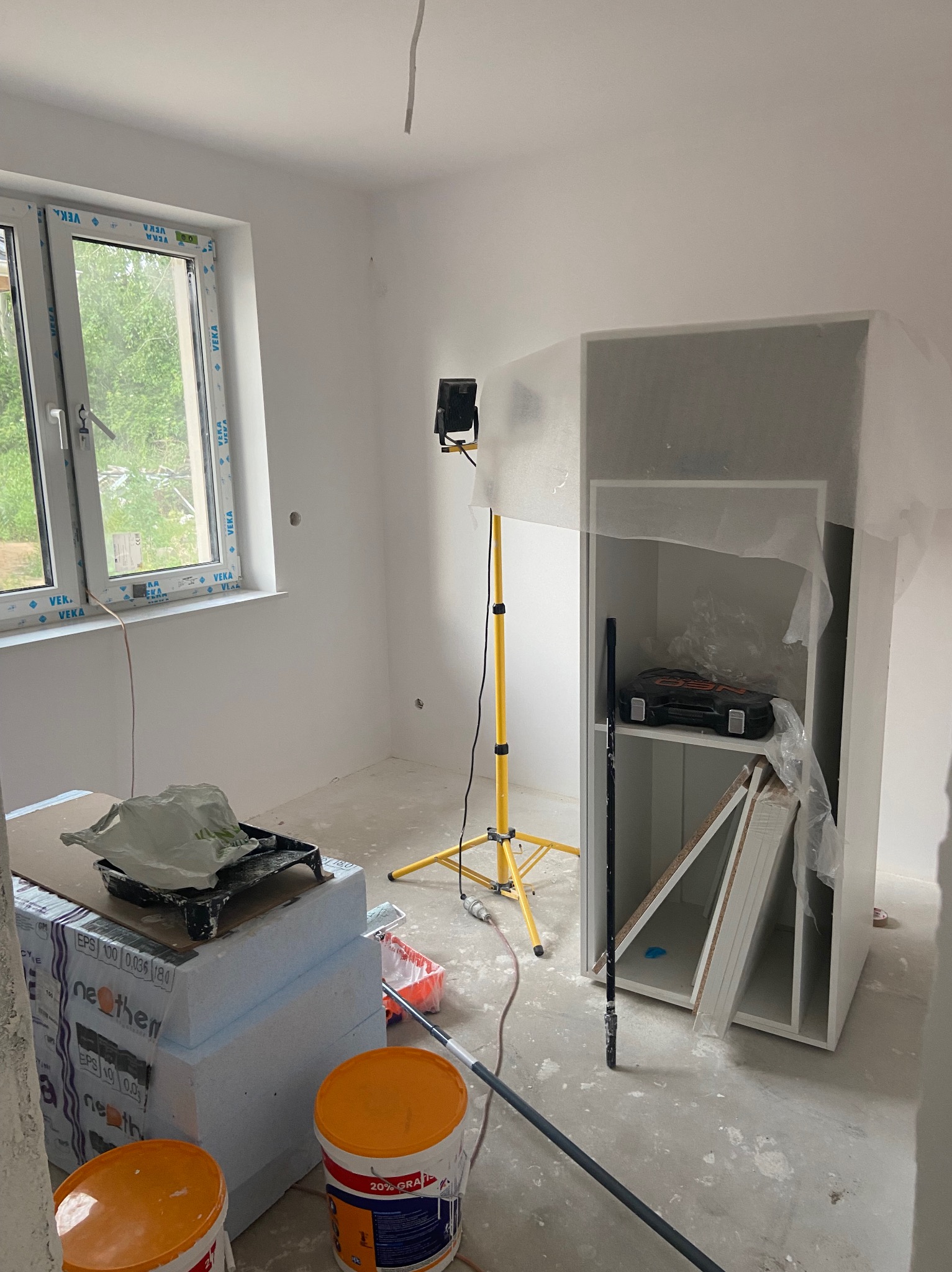 Pokój w trakcie odświeżania: pomalowane ściany na biało, okno z widokiem na zieleń, narzędzia malarskie, lampa na statywie i częściowo zakryty mebel z półkami.