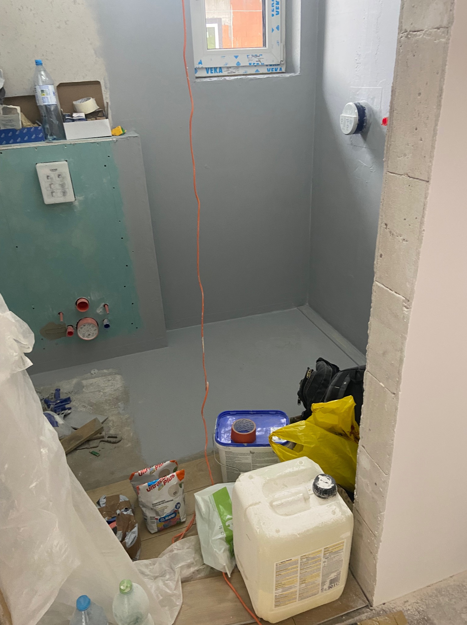 Przygotowana do dalszych prac łazienka: szare ściany, uszczelniona podłoga, instalacja wodna ukryta za płytą gipsową. Widoczne materiały budowlane i narzędzia. Okno VEKA w tle.