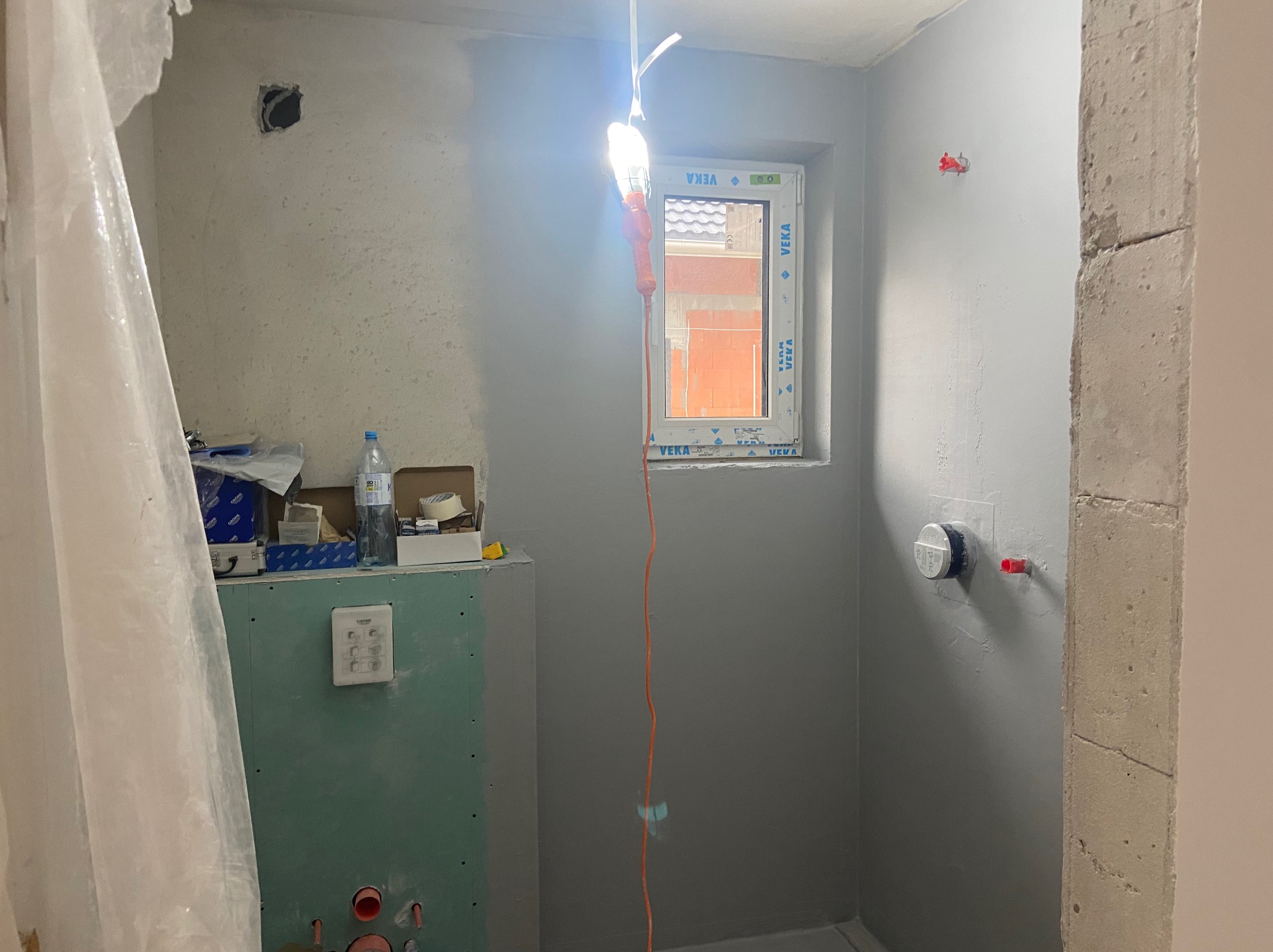 Łazienka w trakcie prac wykończeniowych: szpachlowane ściany, zabudowa z płyty GK, okno z widokiem na ceglany budynek, wisząca lampa robocza.