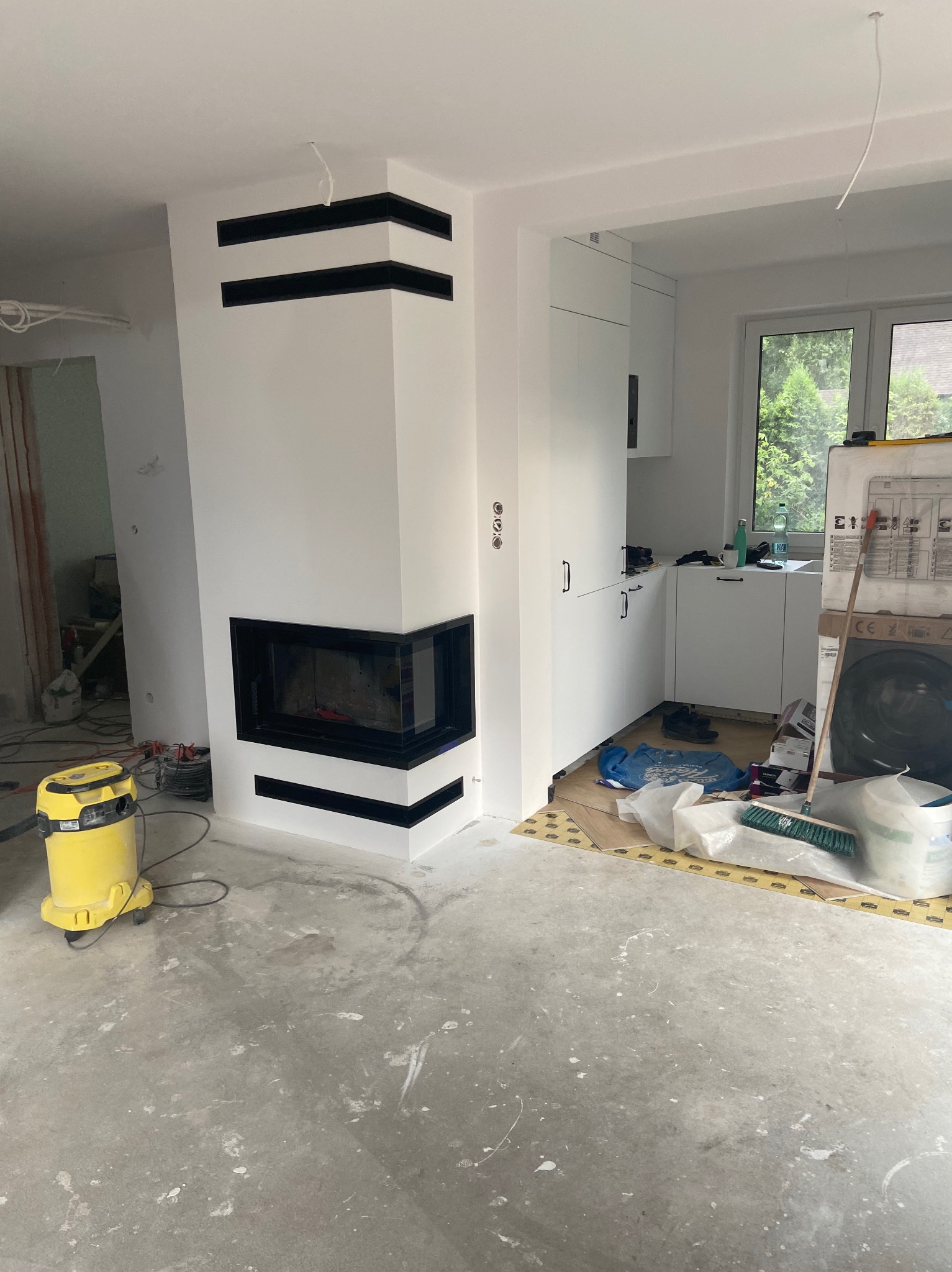 Biały kominek narożny z czarnymi akcentami w surowym wnętrzu. Widoczny odkurzacz, sugerujący prace wykończeniowe. W tle zabudowa kuchenna i okno.