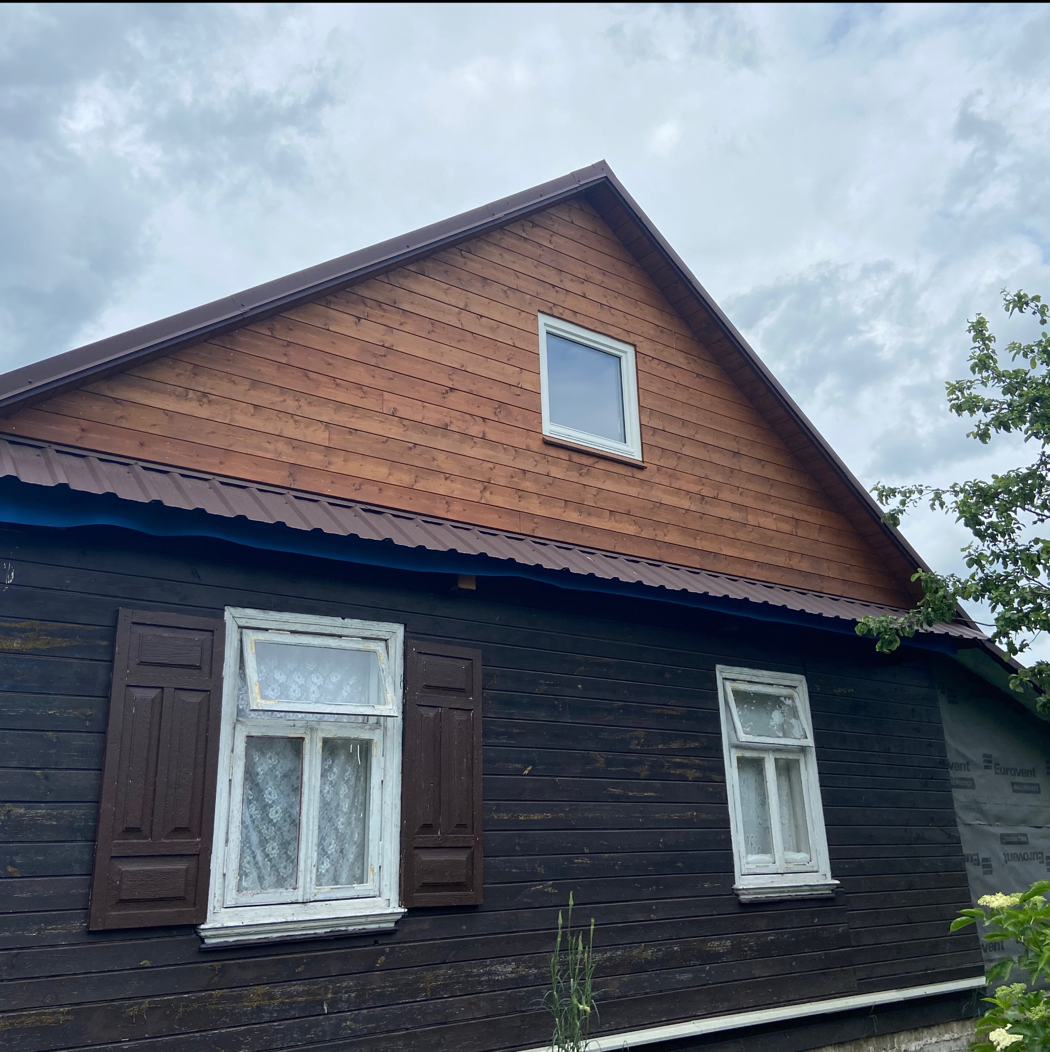 Drewniana elewacja domu z brązowym dachem, białymi oknami i brązowymi okiennicami. Część elewacji odnowiona, część w stanie surowym. Pochmurne niebo w tle.