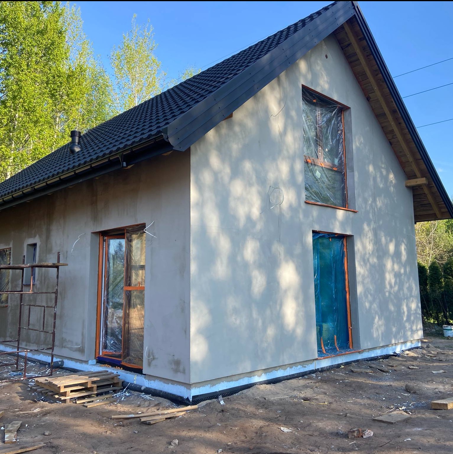 Dom jednorodzinny w trakcie szpachlowania elewacji, okna i drzwi zabezpieczone folią. Widoczne rusztowanie i palety. Tło: zieleń drzew i błękit nieba.