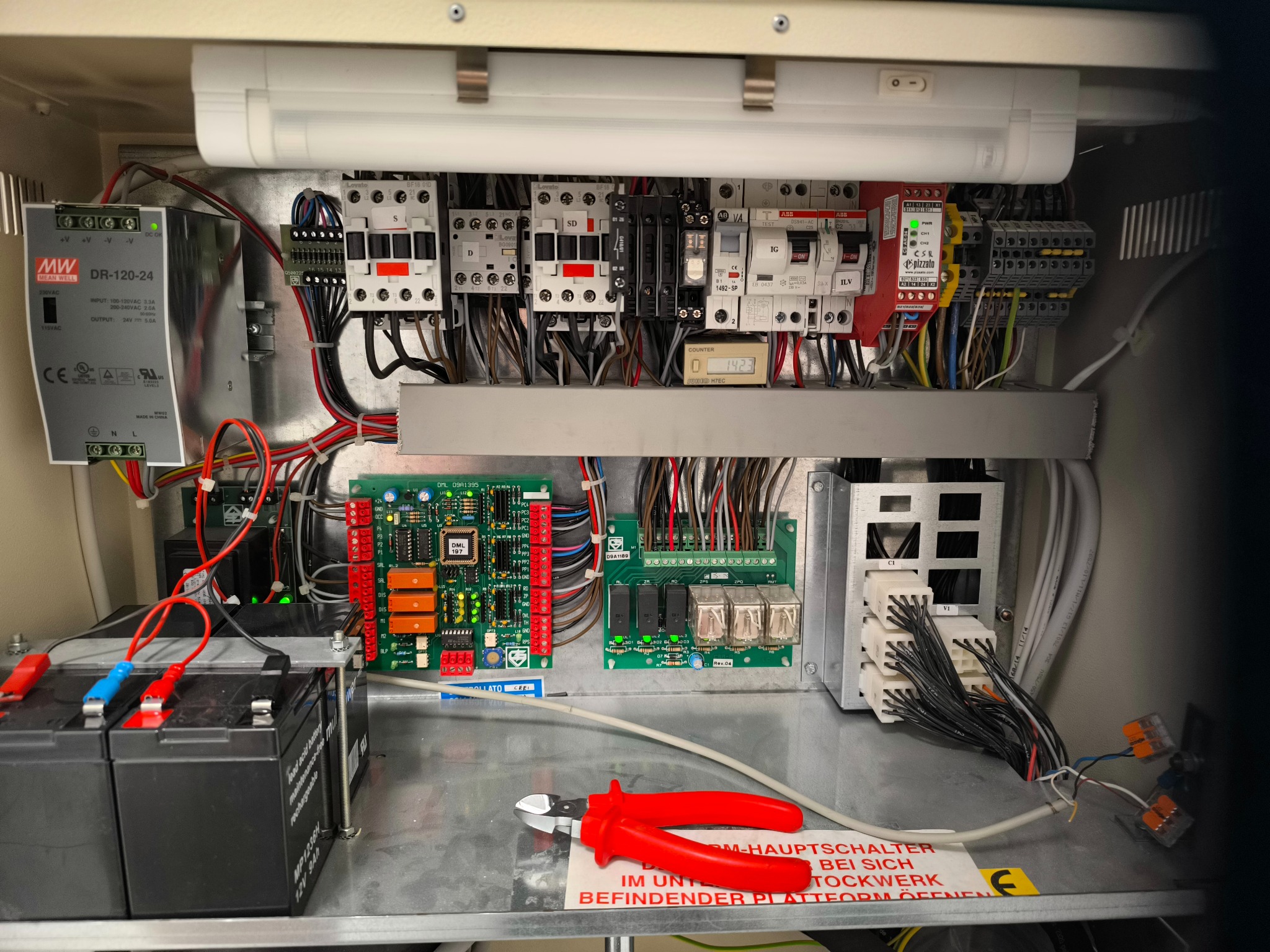 Otwarta szafa sterownicza z panelami elektrycznymi, przewodami, bezpiecznikami i narzędziami, oświetlona lampą LED. Widoczne akumulatory i instrukcja bezpieczeństwa.