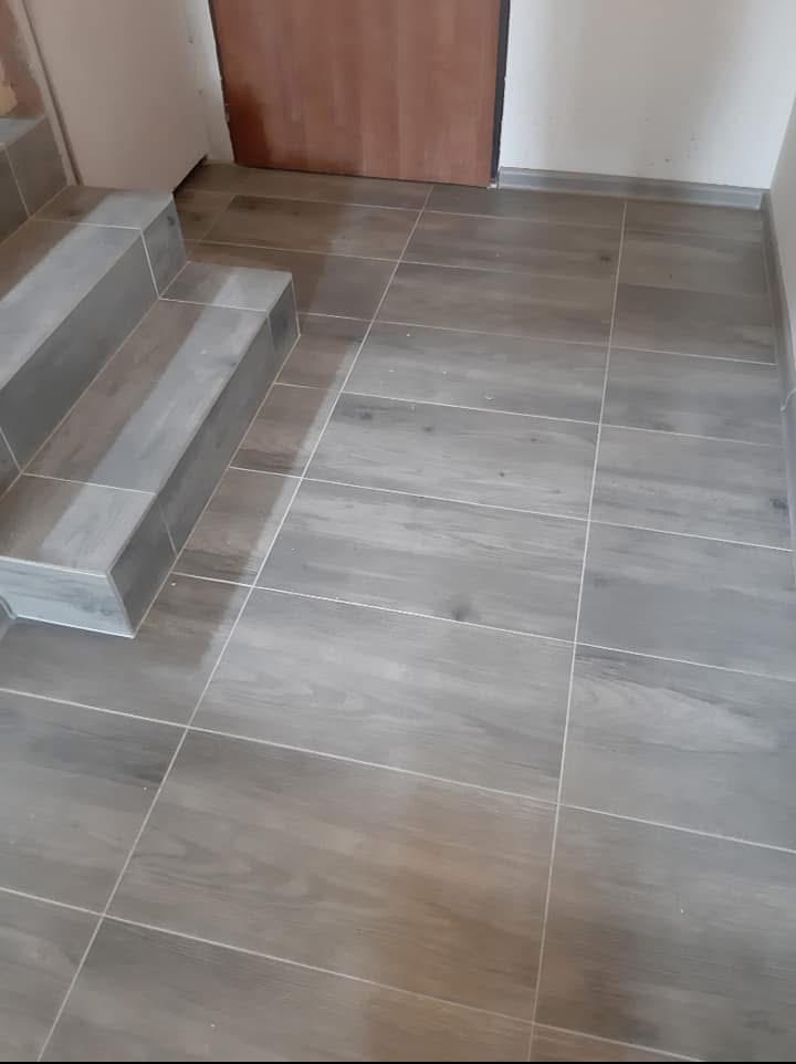 Wykończone schody i podłoga z szarych, drewnopodobnych płytek. Widoczne fugi i listwa przypodłogowa. Ujęcie z bliska, z widoczną fakturą i detalami.