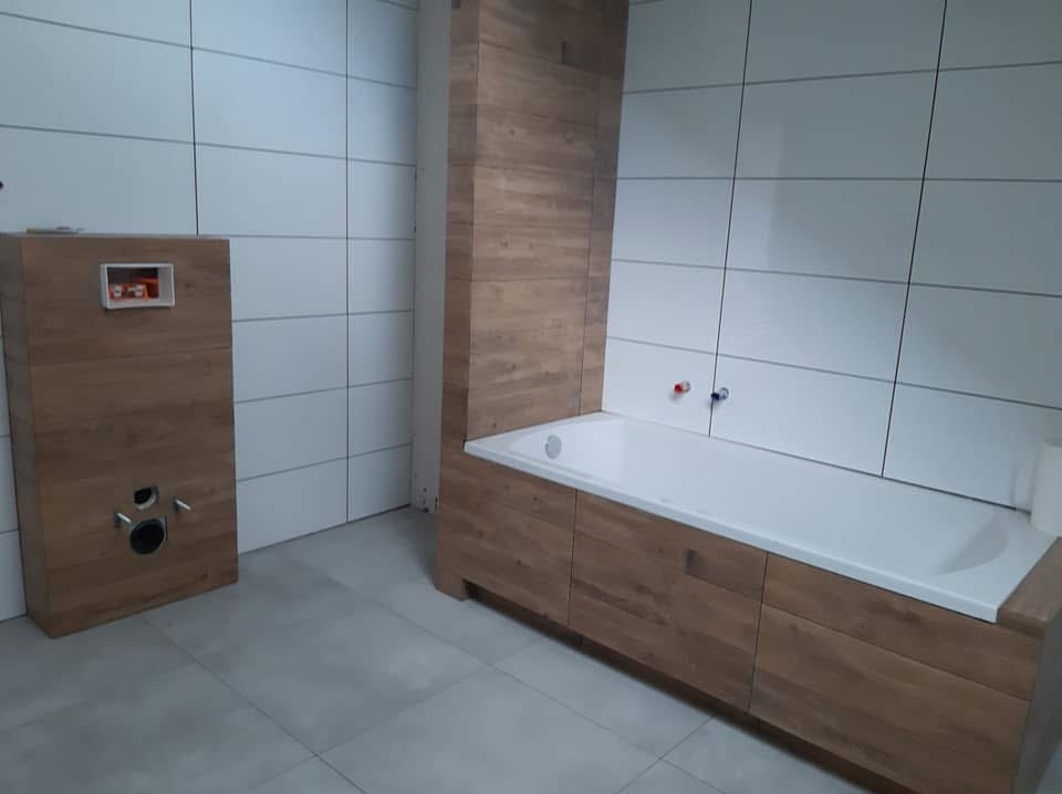 Nowoczesna łazienka w trakcie wykańczania: białe płytki, obudowa wanny i stelaż WC z drewnopodobnego materiału. Widoczne przyłącza wodne.