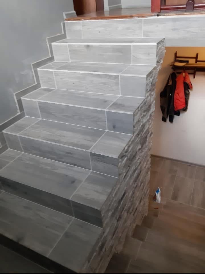 Schody wyłożone szarymi płytkami imitującymi drewno, z dekoracyjnym obłożeniem kamieniem na boku. Widoczny fragment podłogi i wieszak z ubraniami w tle.