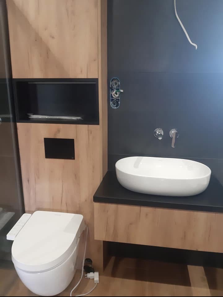 Nowoczesna łazienka z wiszącą miską WC z panelem sterującym, umywalką nablatową i drewnopodobną zabudową. Widoczne niedokończone podłączenie baterii.