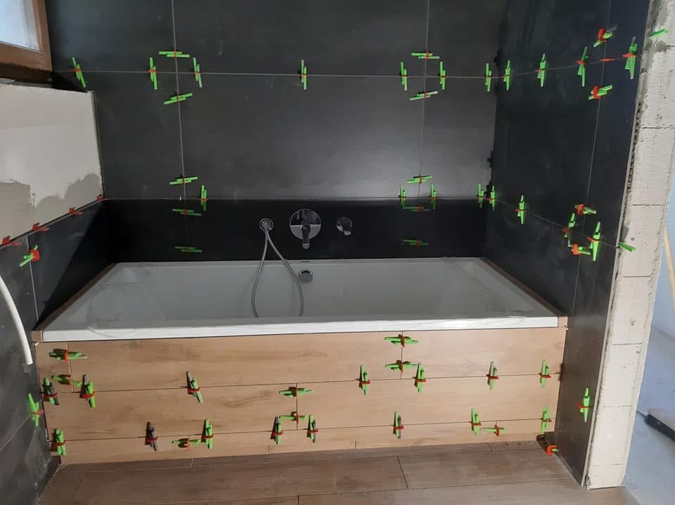 Montaż płytek w łazience: ciemne płytki na ścianach i drewnopodobne na obudowie wanny, widoczne krzyżyki dystansowe i sznurki poziomujące. Prace w toku.