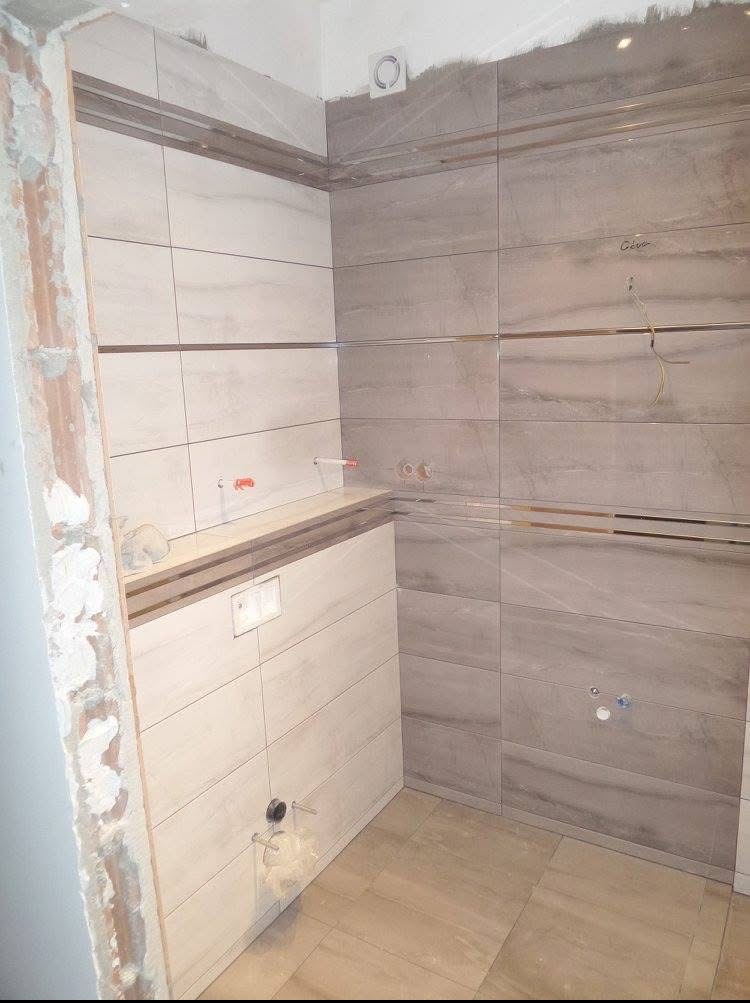 Wykończona łazienka z szarymi i białymi płytkami imitującymi marmur, z widocznymi instalacjami i elementami armatury. Częściowo wykończone rogi ścian.