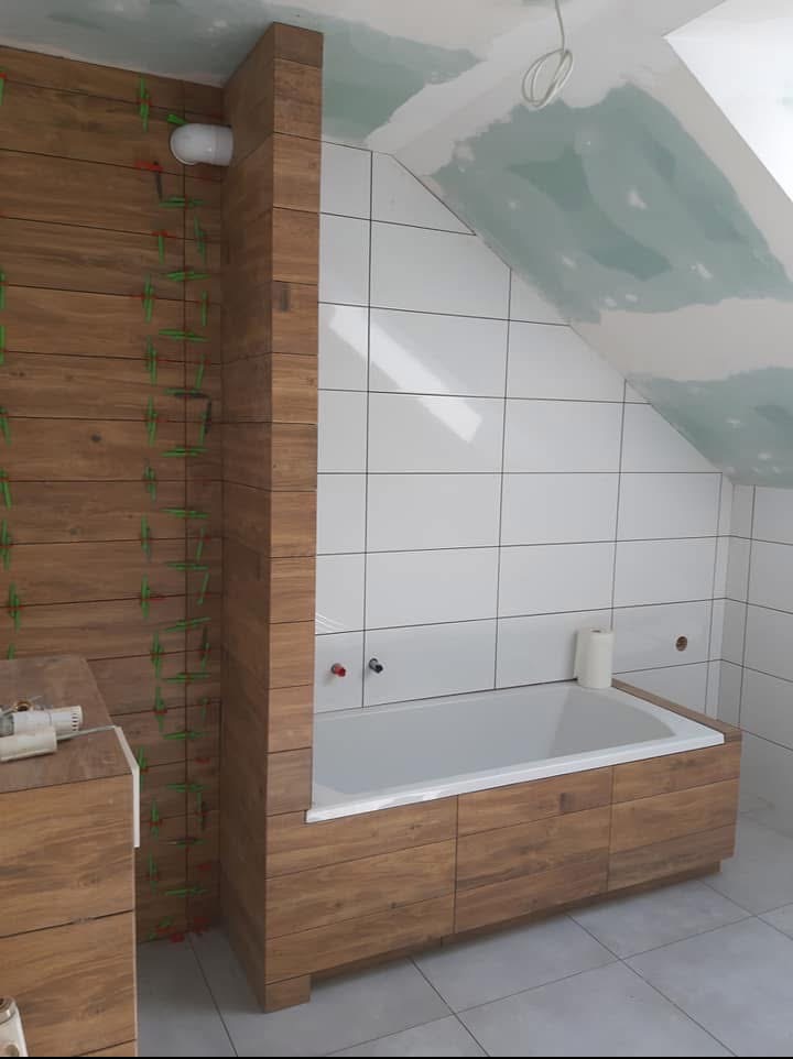 Białe i drewnopodobne płytki w łazience na poddaszu. Częściowo ułożone, z krzyżykami dystansowymi. Wanna obudowana drewnem. Sufit w trakcie wykańczania gładzią.