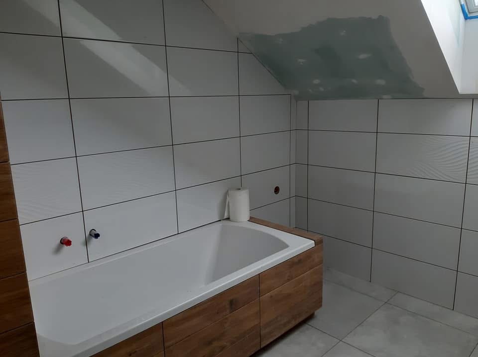 Białe, prostokątne płytki ścienne z ciemnymi fugami w łazience na poddaszu. Obudowana drewnem wanna, częściowo położone płytki na suficie pod skosem.
