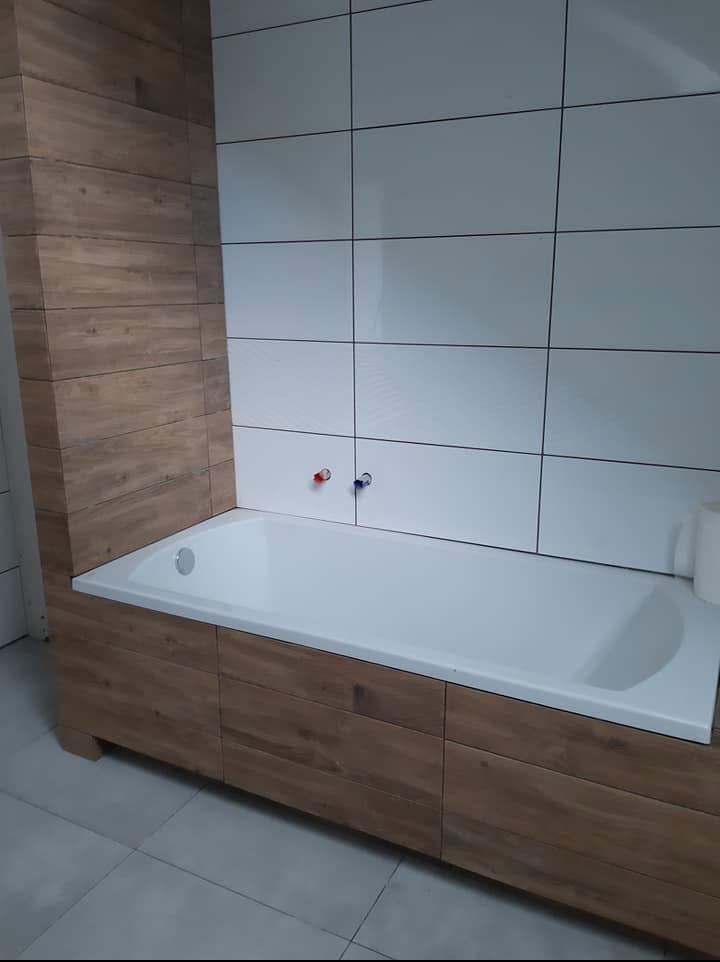 Nowoczesna łazienka z białą wanną obudowaną drewnopodobnymi panelami i białymi, kwadratowymi płytkami na ścianie. Minimalistyczne baterie.