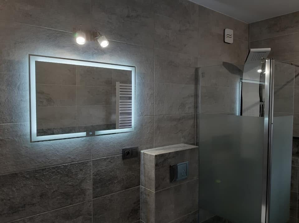 Nowoczesna łazienka z podświetlanym lustrem i szarą glazurą. Widoczny fragment kabiny prysznicowej, oświetlenie punktowe i minimalistyczny design.