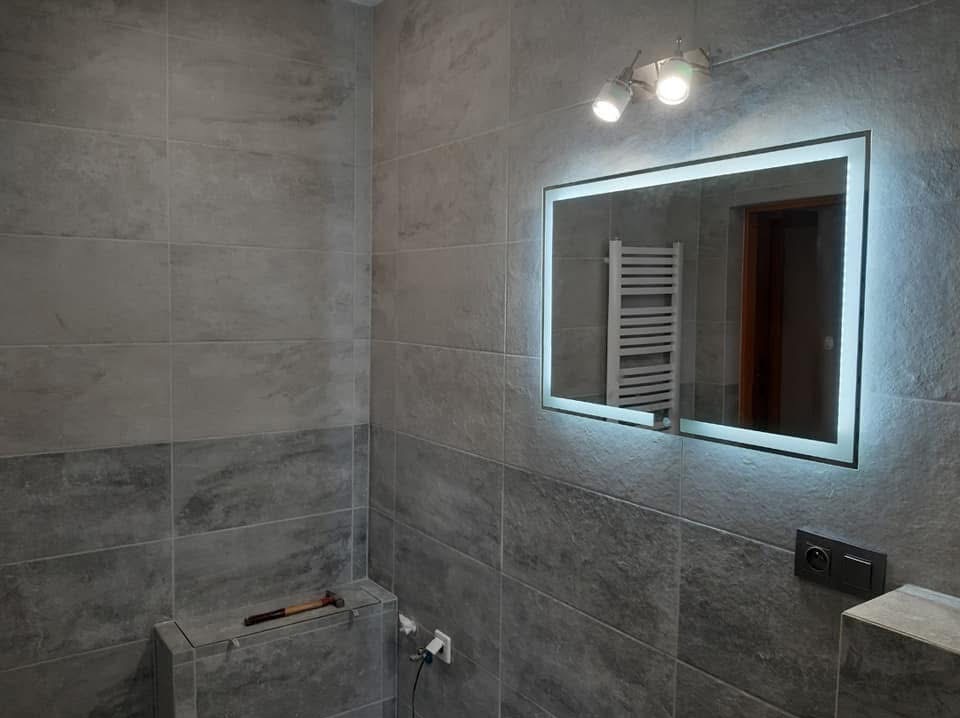 Szara łazienka z podświetlanym lustrem i widocznym w odbiciu grzejnikiem. Na ścianach płytki imitujące kamień, na dole młotek. Nowoczesny design.