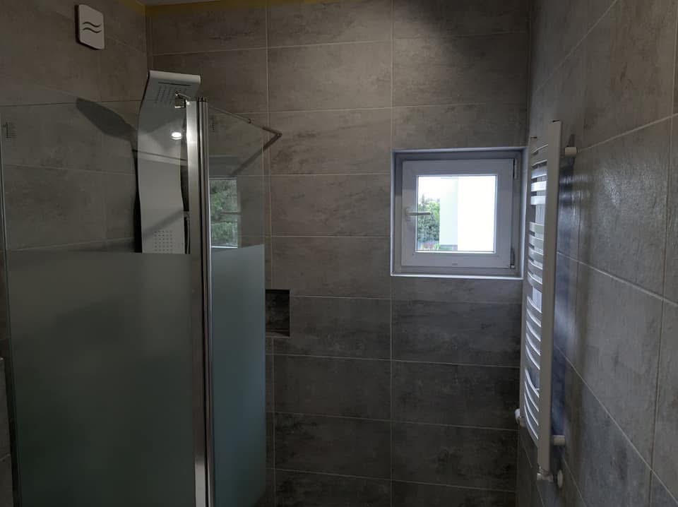 Nowoczesna łazienka z szarymi, podłużnymi płytkami. Kabina prysznicowa z matową szybą i deszczownicą, biały grzejnik drabinkowy i okno z widokiem na zieleń.