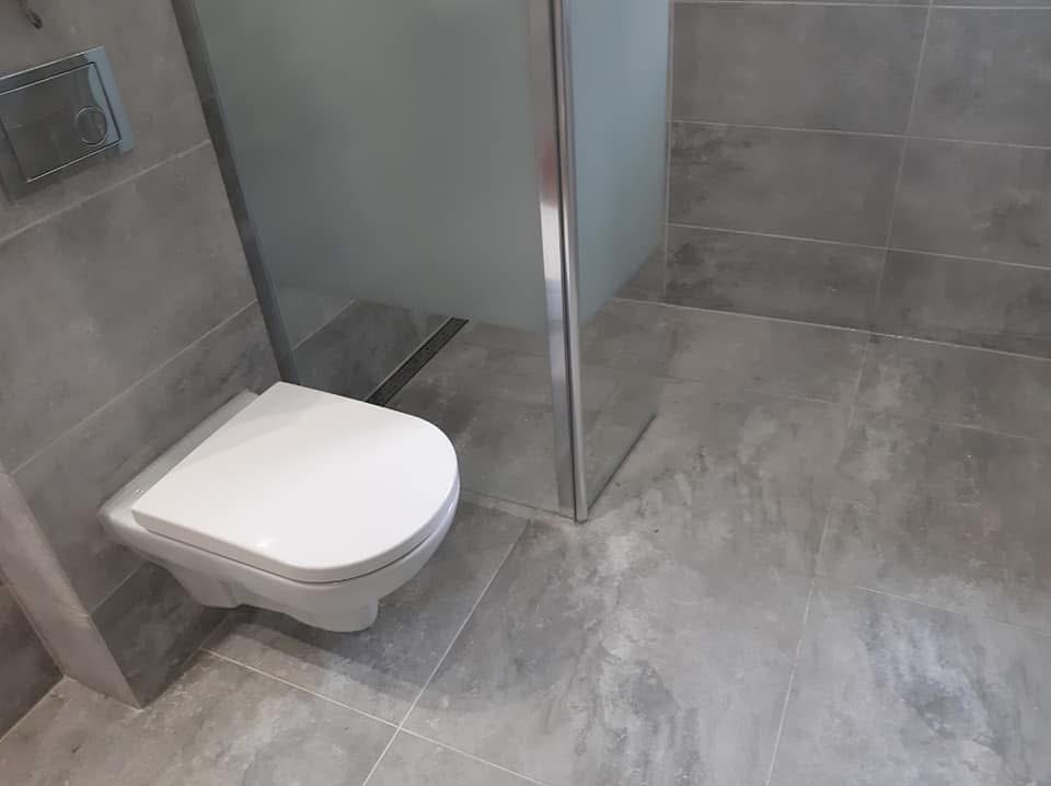 Szara łazienka po remoncie z podwieszaną toaletą i kabiną prysznicową z matowego szkła. Widoczny odpływ liniowy i płytki imitujące beton.