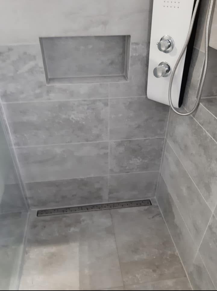 Szare płytki w kabinie prysznicowej z odpływem liniowym i wnęką w ścianie. Nowoczesny panel prysznicowy z chromowanymi pokrętłami.