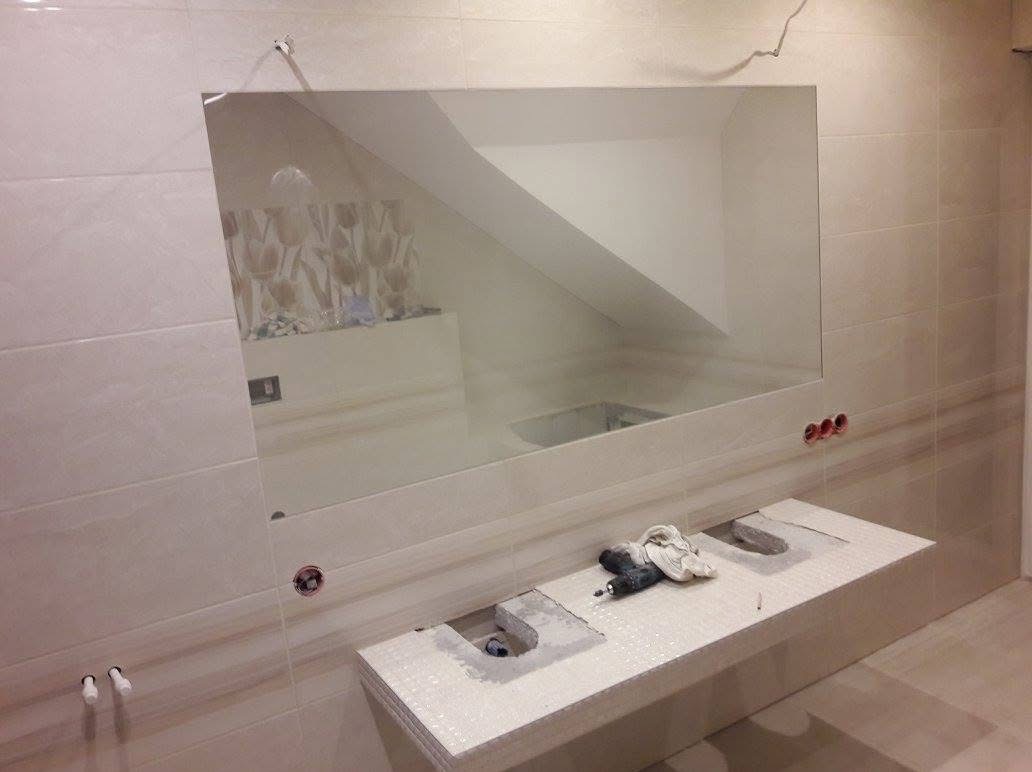 Montaż lustra i umywalki w łazience wykończonej płytkami w odcieniach beżu. Widoczne narzędzia i otwory pod instalacje. Ujęcie z bliska.