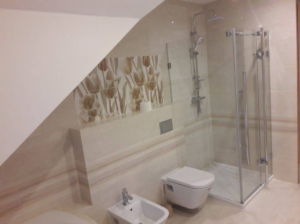 Jasna łazienka po remoncie z kabiną prysznicową, bidetem i toaletą w zabudowie. Na ścianach beżowe płytki z motywem kwiatowym i dekoracyjnymi pasami. Uwagę zwraca ukośny sufit.