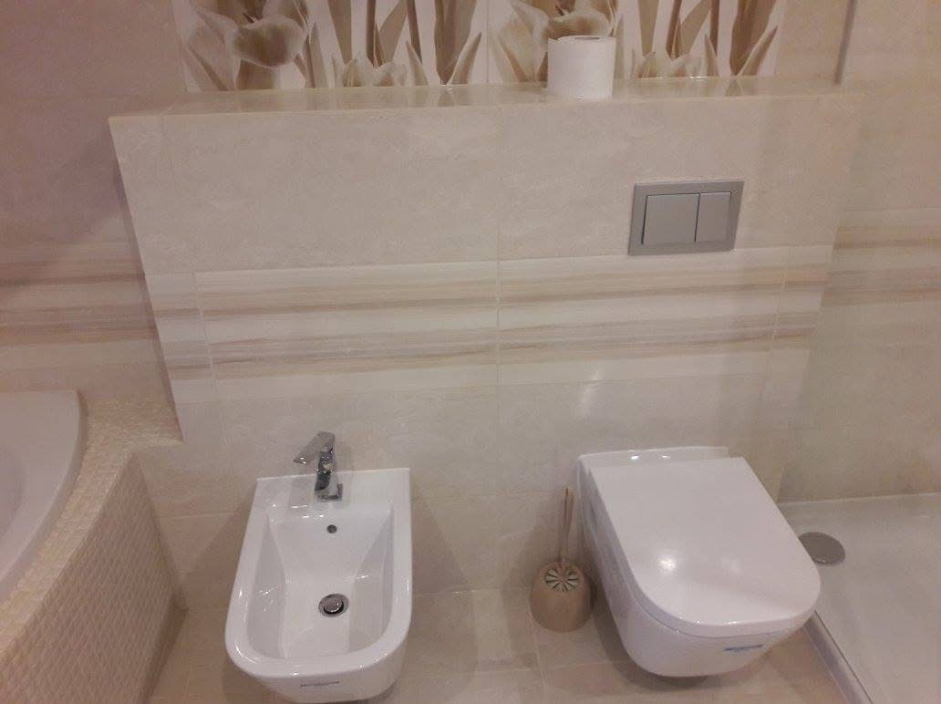 Wykończona łazienka z beżowymi płytkami, wiszącą toaletą i bidetem. Na ścianie widoczny wzór kwiatowy. Całość utrzymana w jasnej tonacji.