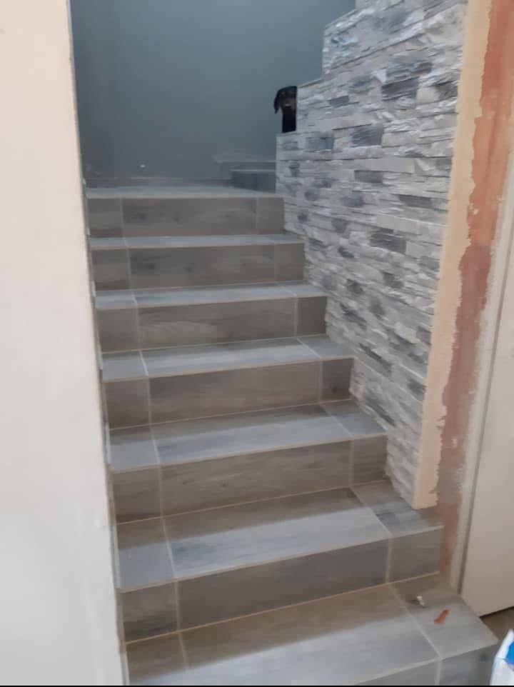 Wykończone schody z szarych płytek, ściana obłożona dekoracyjnym kamieniem. U góry schodów widoczny fragment czarnego psa. Wykończenie w szarościach.