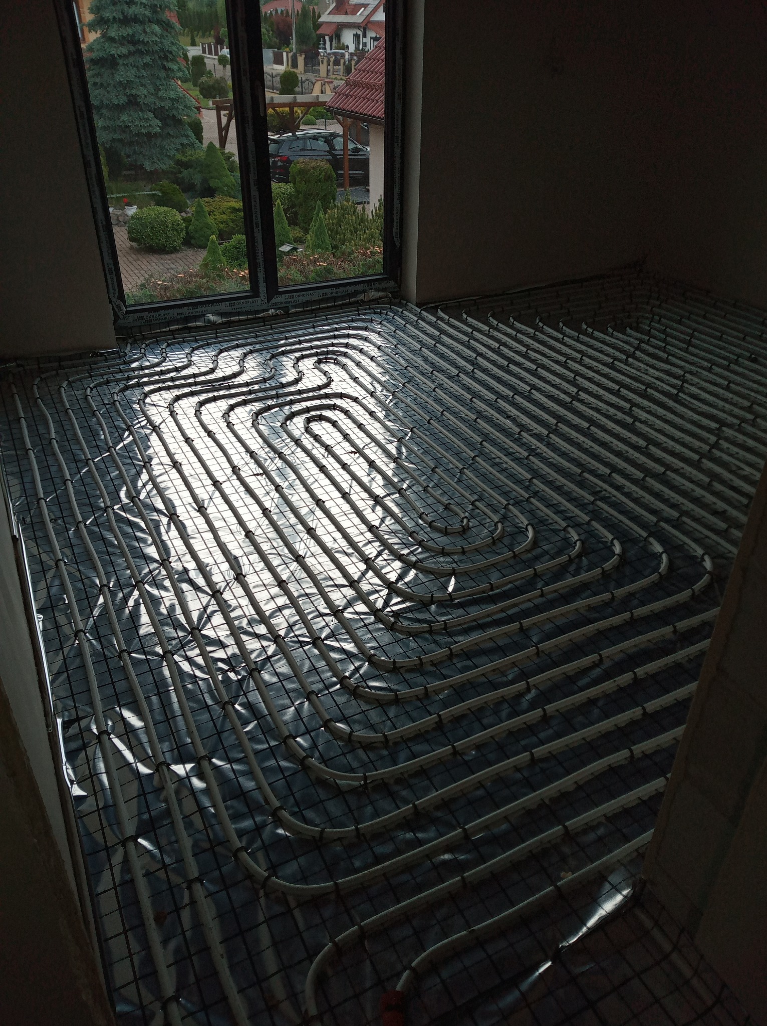 Białe rurki instalacji grzewczej ułożone w pętle na folii pod oknem, przygotowane do zalania posadzką. Widok na ogród przez okno.