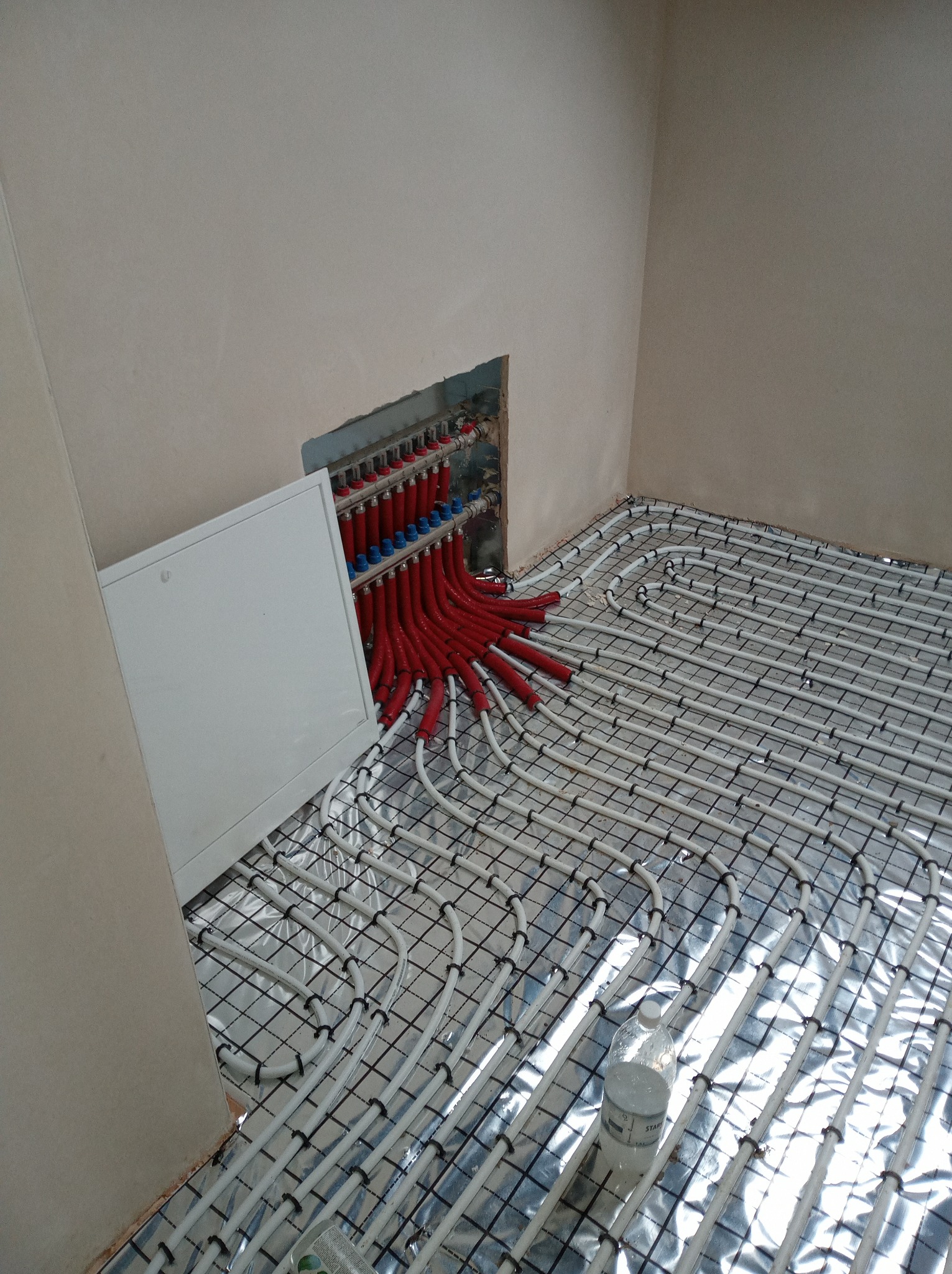 Instalacja rur ogrzewania podłogowego na folii termoizolacyjnej, widoczny rozdzielacz z czerwonymi i niebieskimi rurami. Etap prac przed wylewką.
