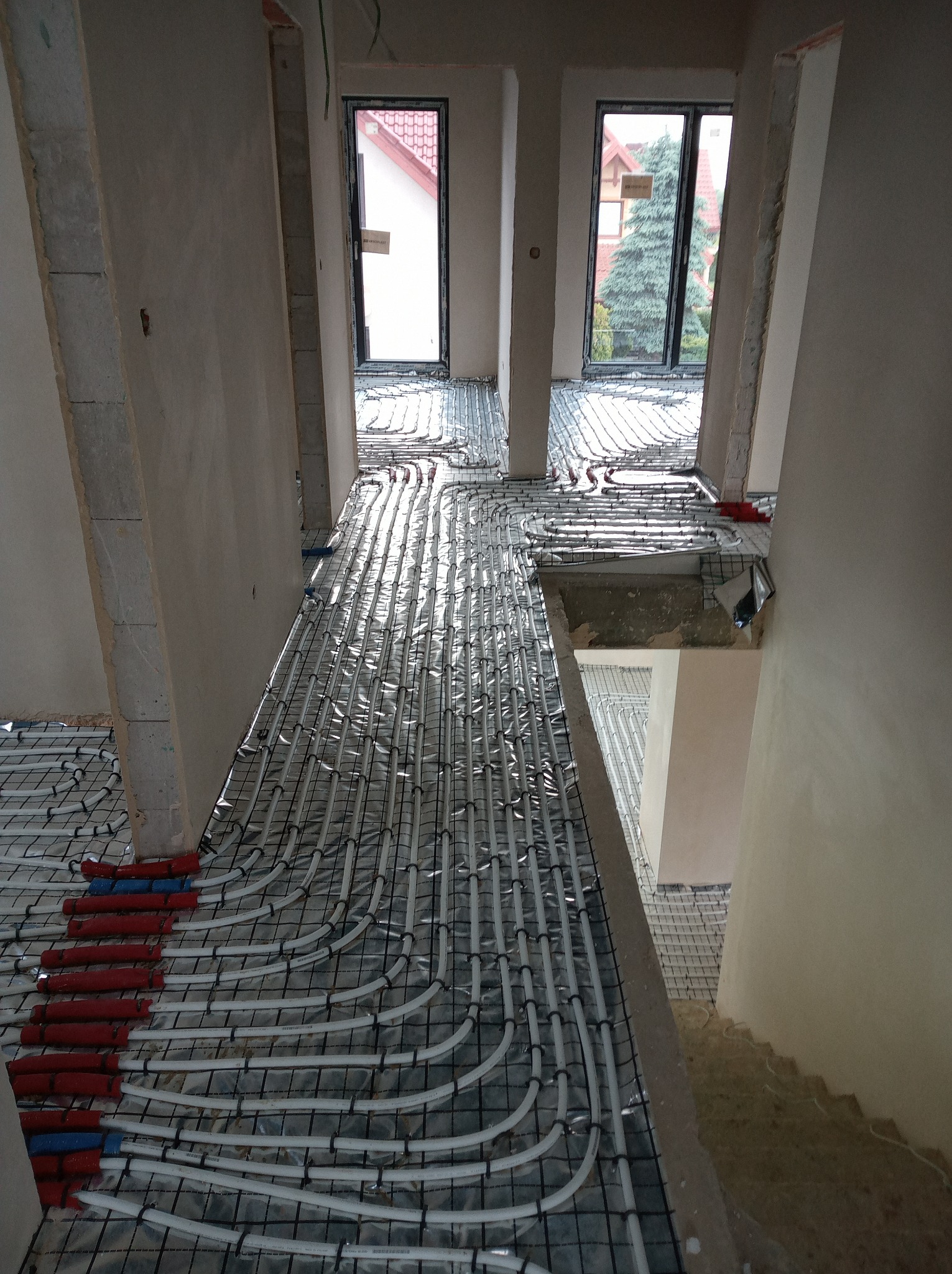 Rozłożone białe rury na folii i siatce w surowym wnętrzu przed wylaniem posadzki. Widoczne okna i fragmenty ścian.