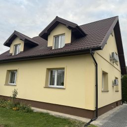 GP-BUD - Żółty dom jednorodzinny z brązowym dachem i dwoma klimatyzatorami na elewacji bocznej, otoczony zielenią i kwiatami. Widok z perspektywy ogrodu.