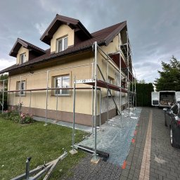 GP-BUD - Dom jednorodzinny w trakcie prac elewacyjnych, widoczne rusztowania, przygotowane podłoże pod ocieplenie. Na podjeździe zaparkowany samochód osobowy i bus.