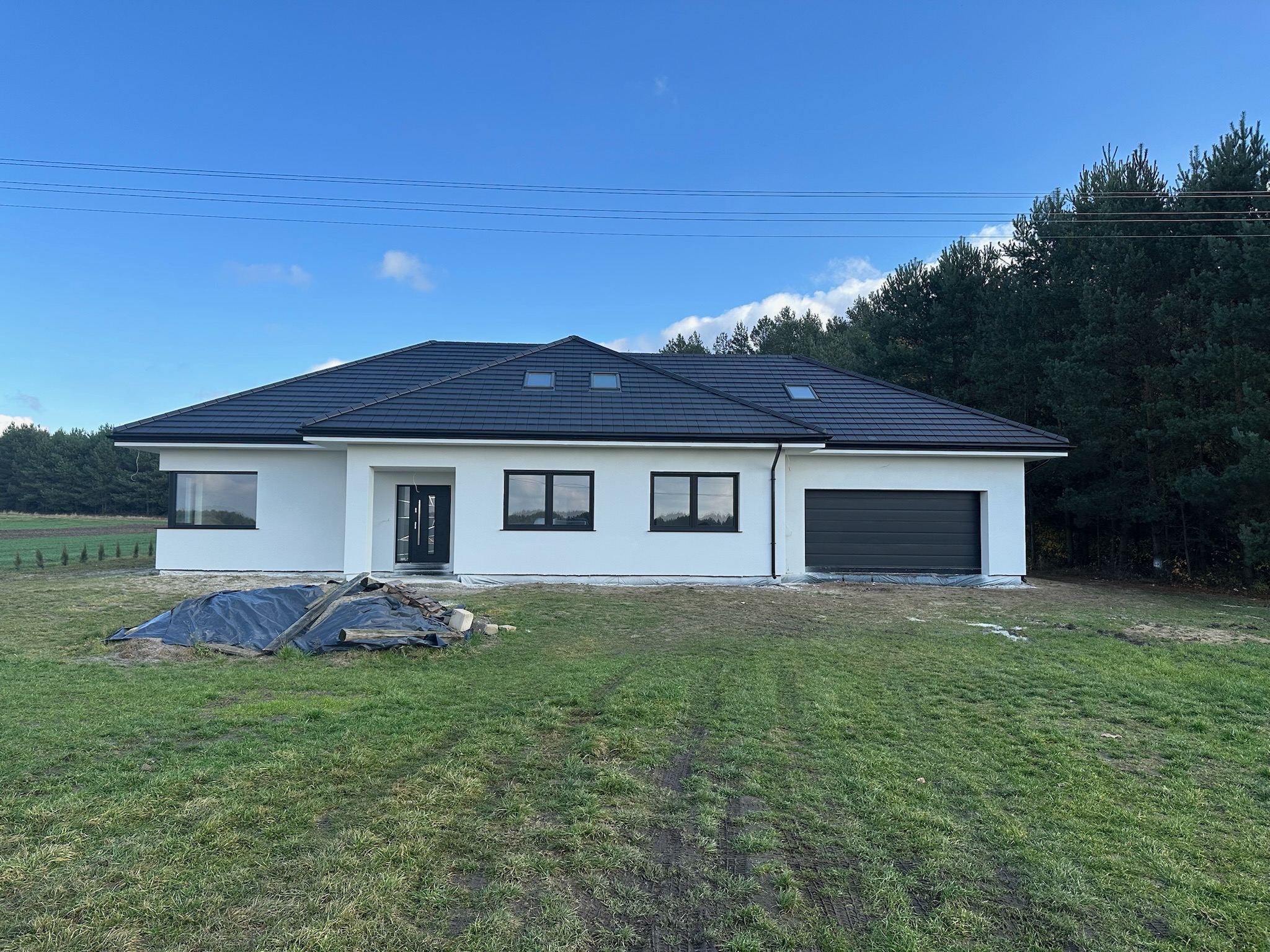 Nowoczesny, parterowy dom z grafitowym dachem i garażem, białą elewacją oraz dużymi oknami, na tle zielonego trawnika i lasu. Widoczne pozostałości po budowie.