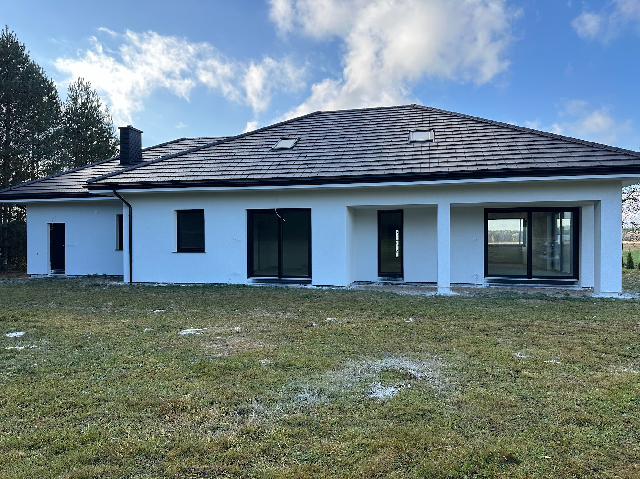 Nowoczesny, parterowy dom z białymi ścianami i ciemną dachówką, widoczny z perspektywy ogrodu w słoneczny dzień. Minimalistyczna architektura z dużymi przeszkleniami.