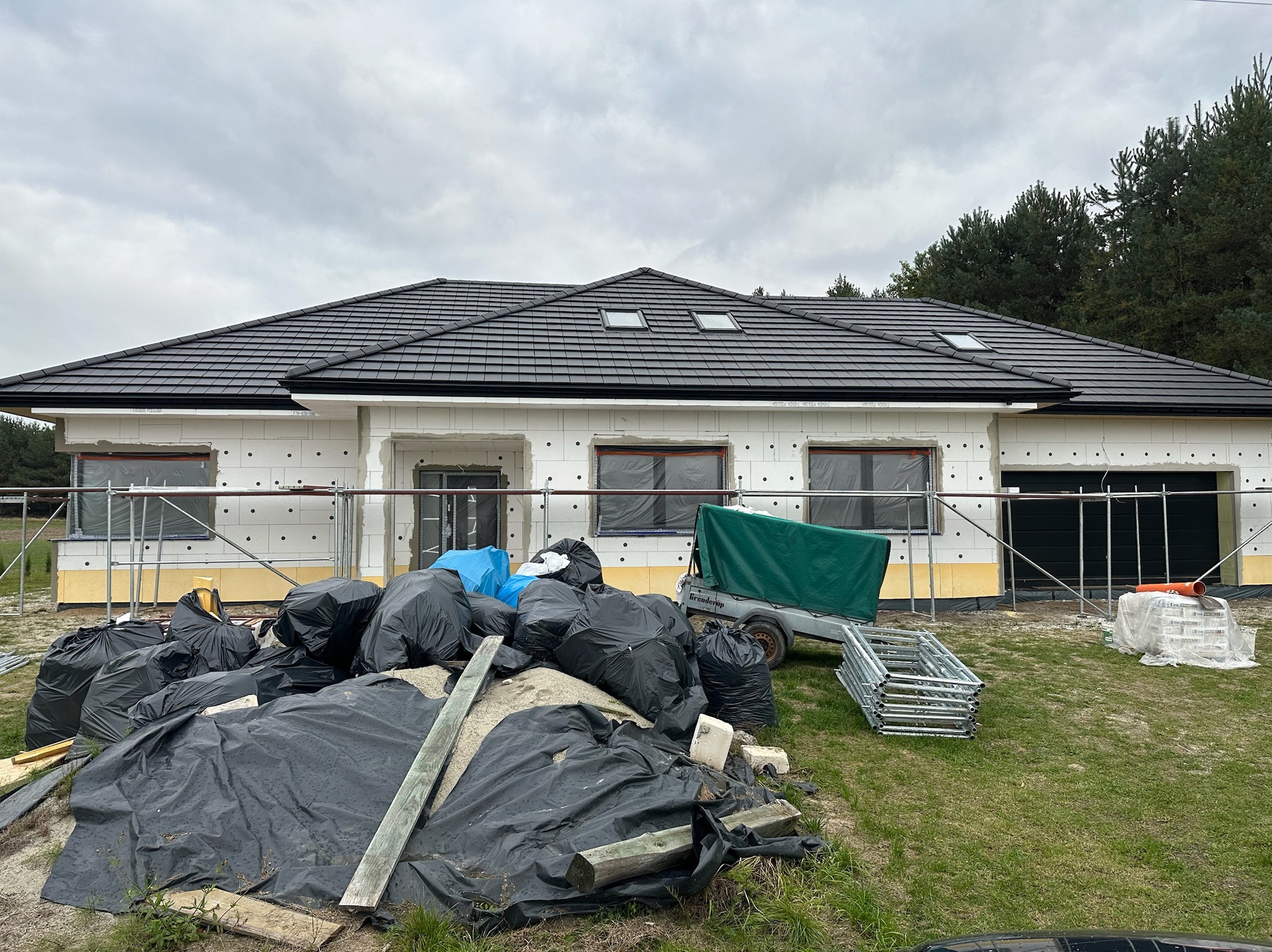 Dom jednorodzinny w trakcie ocieplania, widoczne worki z odpadami budowlanymi, rusztowanie i przyczepka. Elewacja z białych bloczków, czarny dach z oknami.