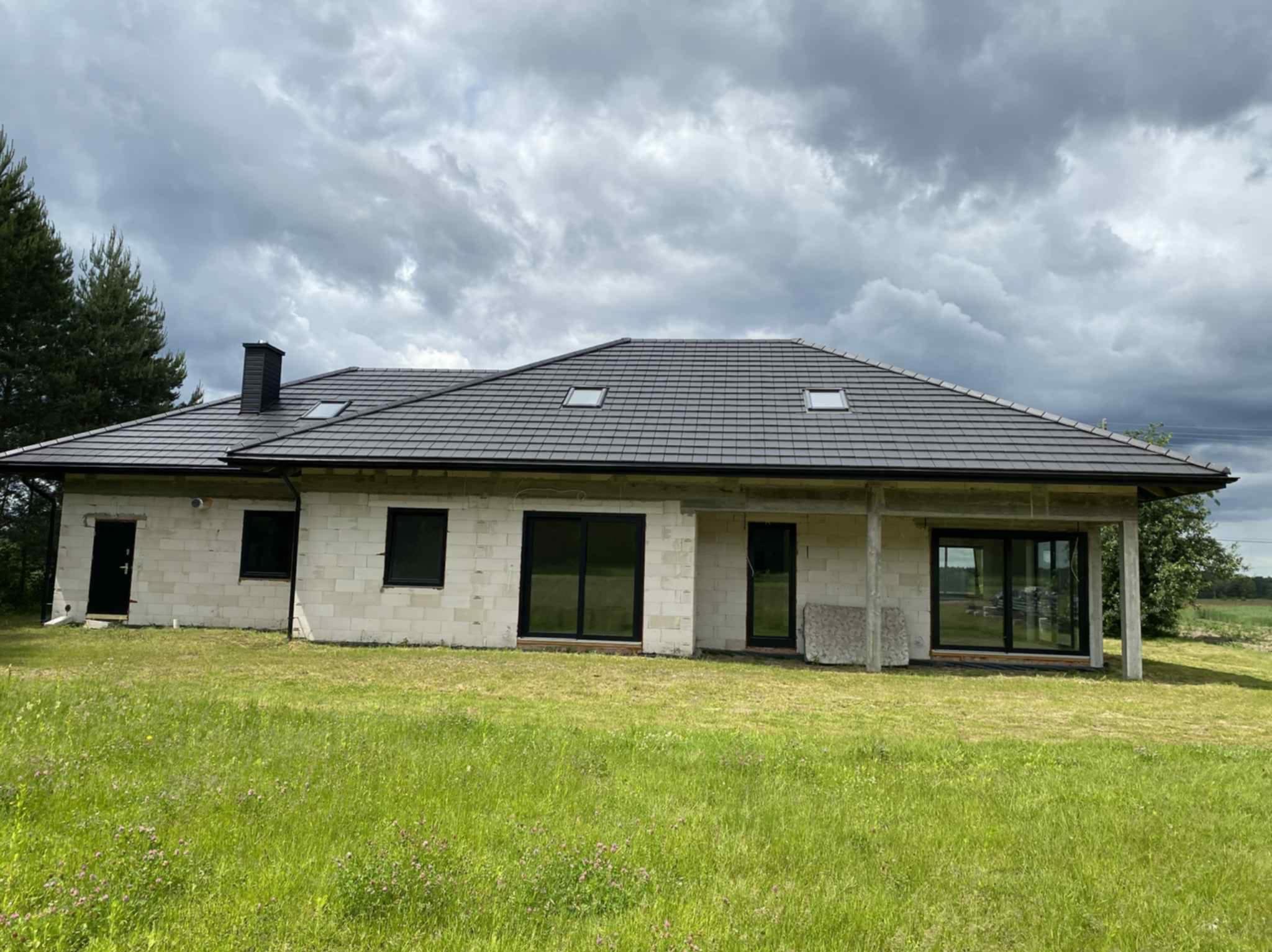 Surowy dom z czarnym dachem w Zamościu, widok z zewnątrz. Budynek w trakcie budowy, z widocznymi pustakami i oknami. Pochmurne niebo w tle.