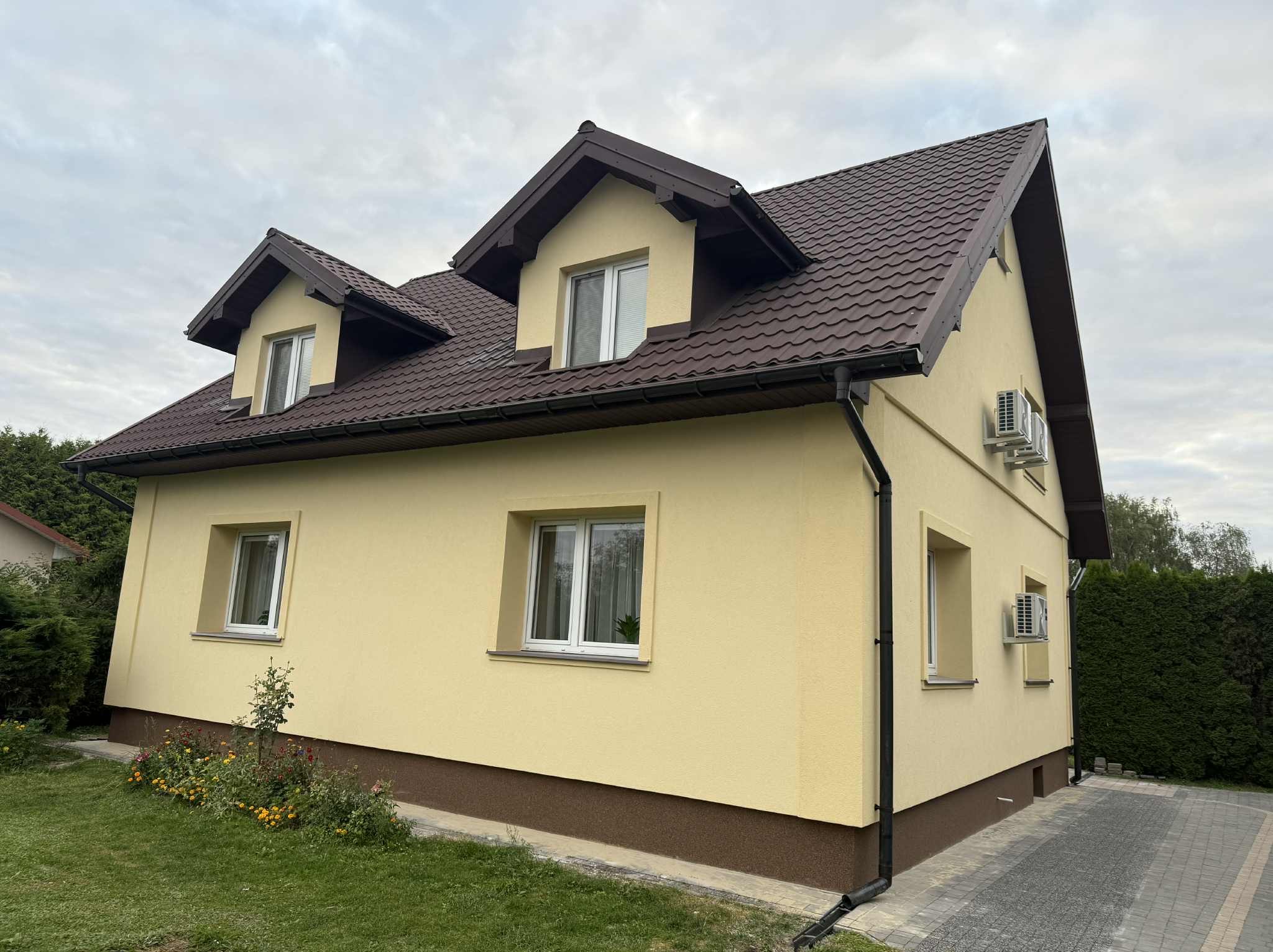 Żółty dom jednorodzinny z brązowym dachem i dwoma klimatyzatorami na elewacji bocznej, otoczony zielenią i kwiatami. Widok z perspektywy ogrodu.