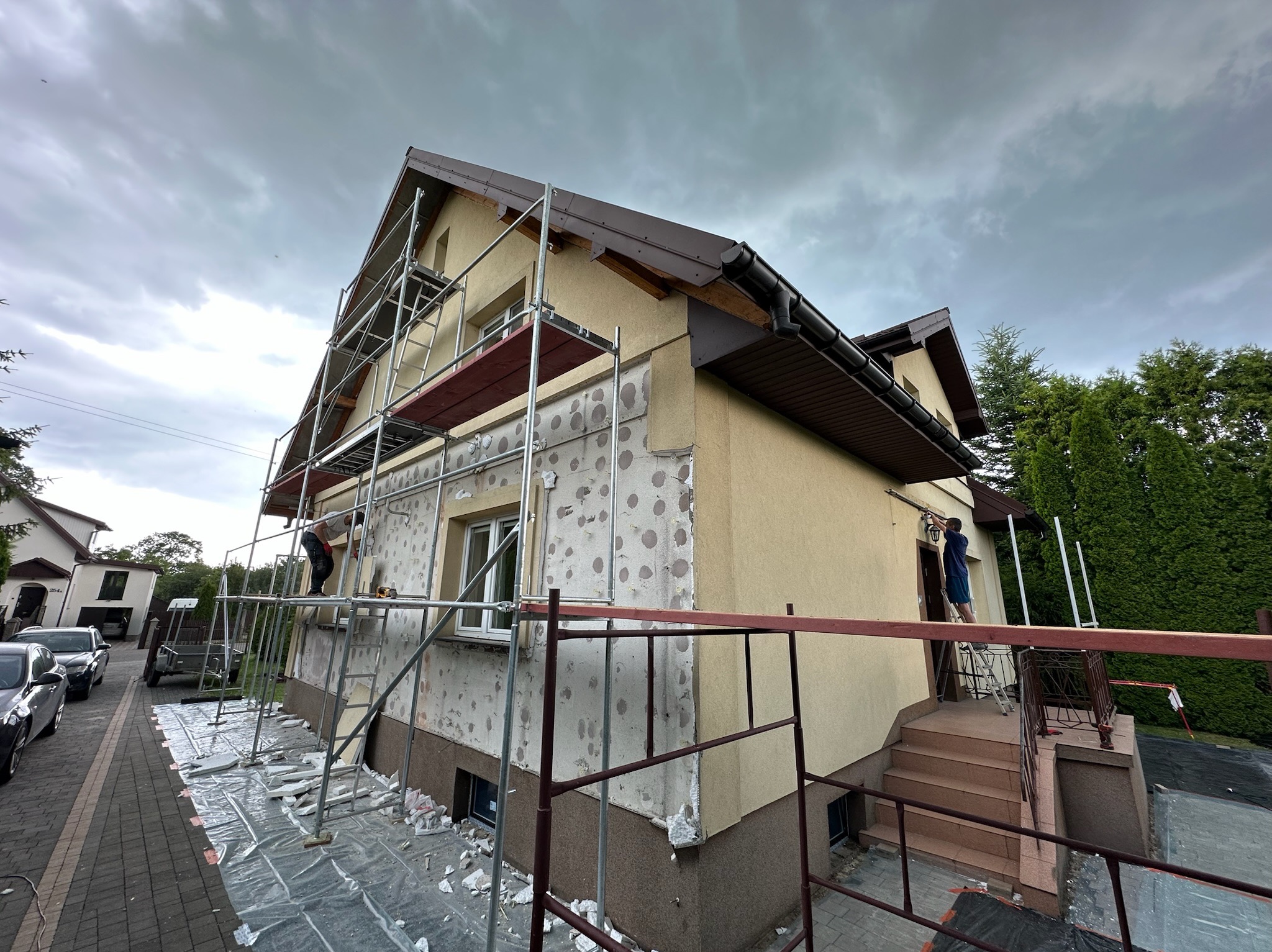 Dom jednorodzinny w trakcie prac elewacyjnych: widoczne rusztowania, fragmenty ocieplenia i pracownicy. Część elewacji przygotowana do dalszych etapów prac.