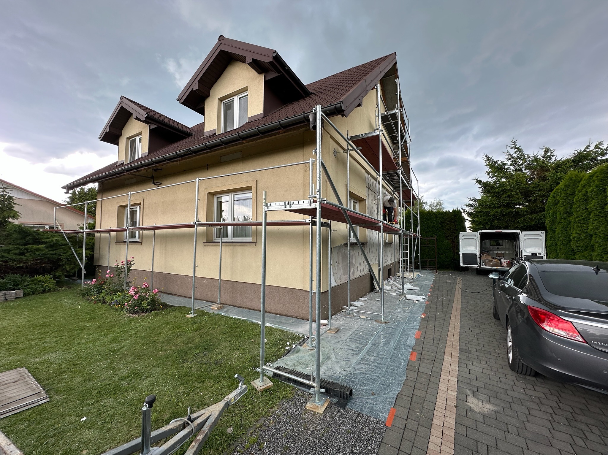 Dom jednorodzinny w trakcie prac elewacyjnych, widoczne rusztowania, przygotowane podłoże pod ocieplenie. Na podjeździe zaparkowany samochód osobowy i bus.