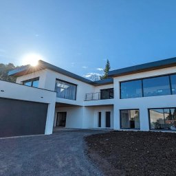 Nova House Poland Sp. z o.o. - Nowoczesny, dwukondygnacyjny dom z płaskim dachem i dużymi oknami, garaż z szarą bramą, widok z dołu na tle błękitnego nieba z promieniami słońca.