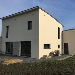 Nova House Poland Sp. z o.o. - Nowoczesny, dwukondygnacyjny dom z jasną elewacją i grafitowymi oknami. Widoczny fragment tarasu z meblami i świeżo posadzona trawa. Minimalistyczny design.