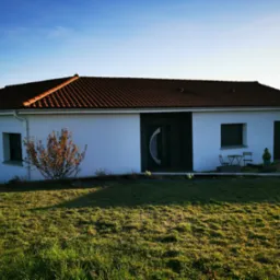 Parterowy, biały dom z brązowym dachem w słoneczny dzień. Widoczne okna, drzwi z elementami szkła, trawnik i niewielki ogródek przed domem. Minimalistyczny styl.