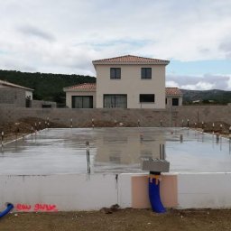 Nova House Poland Sp. z o.o. - Wylana płyta fundamentowa pod budowę domu jednorodzinnego. Widoczne zbrojenia, instalacje i bloczki fundamentowe. W tle budynki w trakcie budowy i krajobraz.