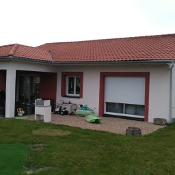 Nova House Poland Sp. z o.o. - Parterowy dom z czerwonym dachem i obramowaniami okien. Na posesji trawnik i zabawki dziecięce. Widoczny fragment drewnianego placu zabaw.