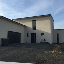 Nova House Poland Sp. z o.o. - Nowoczesny dom z garażem i okrągłym oknem, widok z zewnątrz. Na podjeździe żwir i taczka. Minimalistyczna architektura, jasna elewacja.