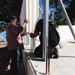 Nova House Poland Sp. z o.o. - Montaż białych paneli izolacyjnych na fundamencie z wystającymi prętami zbrojeniowymi. Dwóch robotników przy pracy, widok z bliska.