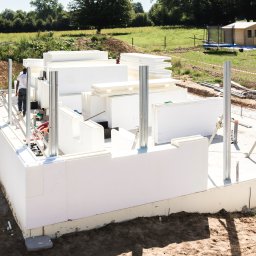 Nova House Poland Sp. z o.o. - Budowa domu z białych paneli izolacyjnych, widoczne metalowe podpory i pracownicy. W tle zielone pole i mały drewniany budynek.