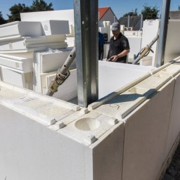 Nova House Poland Sp. z o.o. - Wznoszenie ściany z białych bloczków styropianowych, z widocznymi otworami, wspartych metalowymi stemplami. W tle pracownik w czarnej koszulce i czapce.