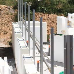 Nova House Poland Sp. z o.o. - Budowa domu modułowego we Wrocławiu: białe ściany z izolacją, stalowe wzmocnienia i zielony wąż na placu budowy. Widok z góry na fundamenty.