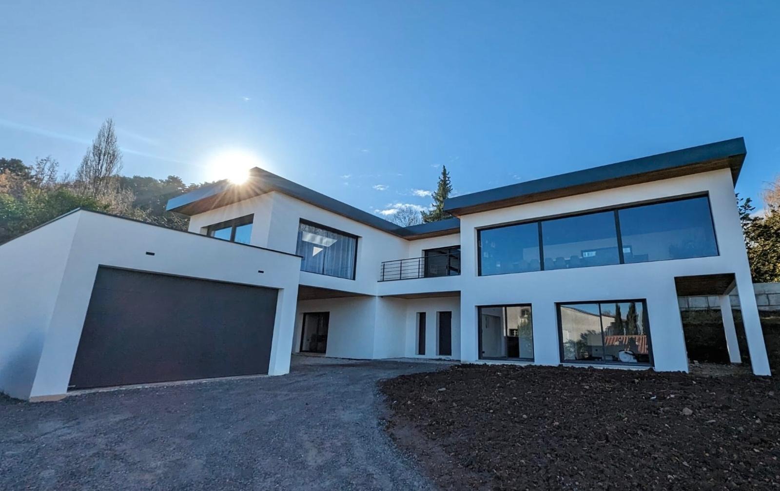 Nowoczesny, dwukondygnacyjny dom z płaskim dachem i dużymi oknami, garaż z szarą bramą, widok z dołu na tle błękitnego nieba z promieniami słońca.